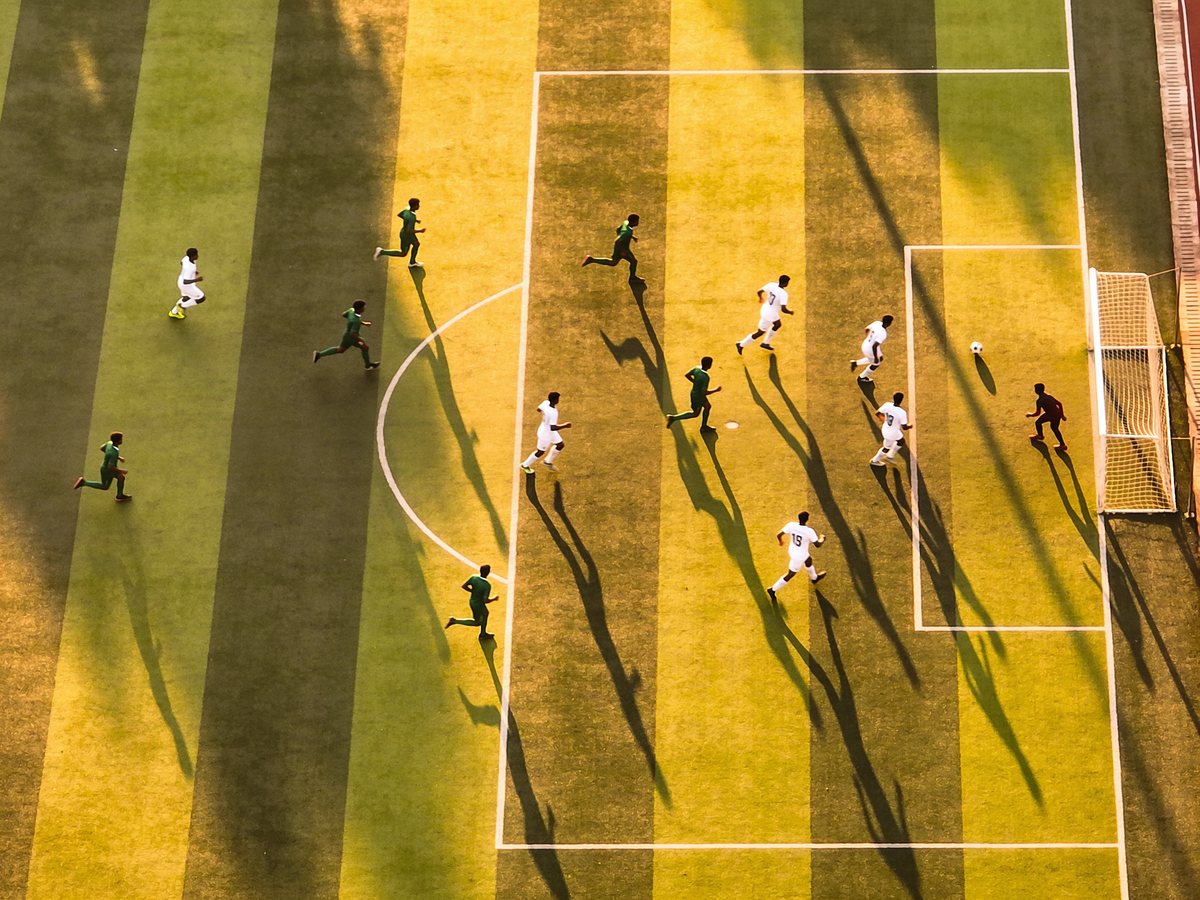 A football match seen from straight above at golden hour, every player and every long shadow visible in one attacking move