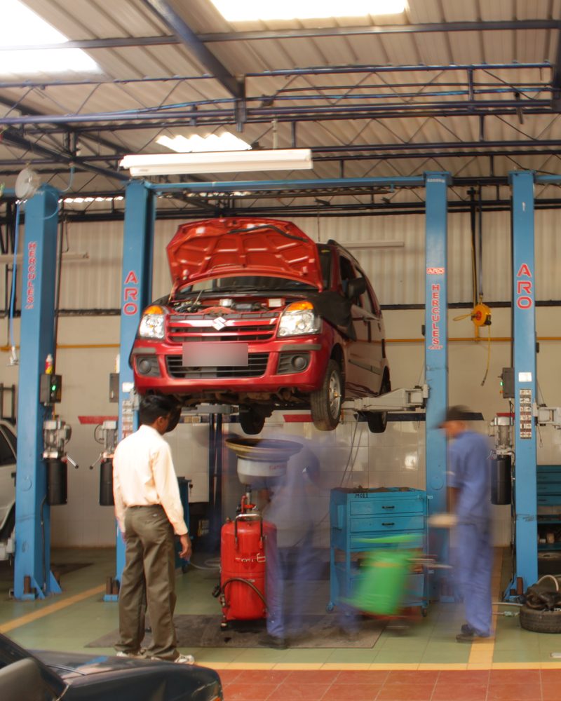 A car raised on a lift in a Bangalore service station, a man in a shirt watching as mechanics hurry past